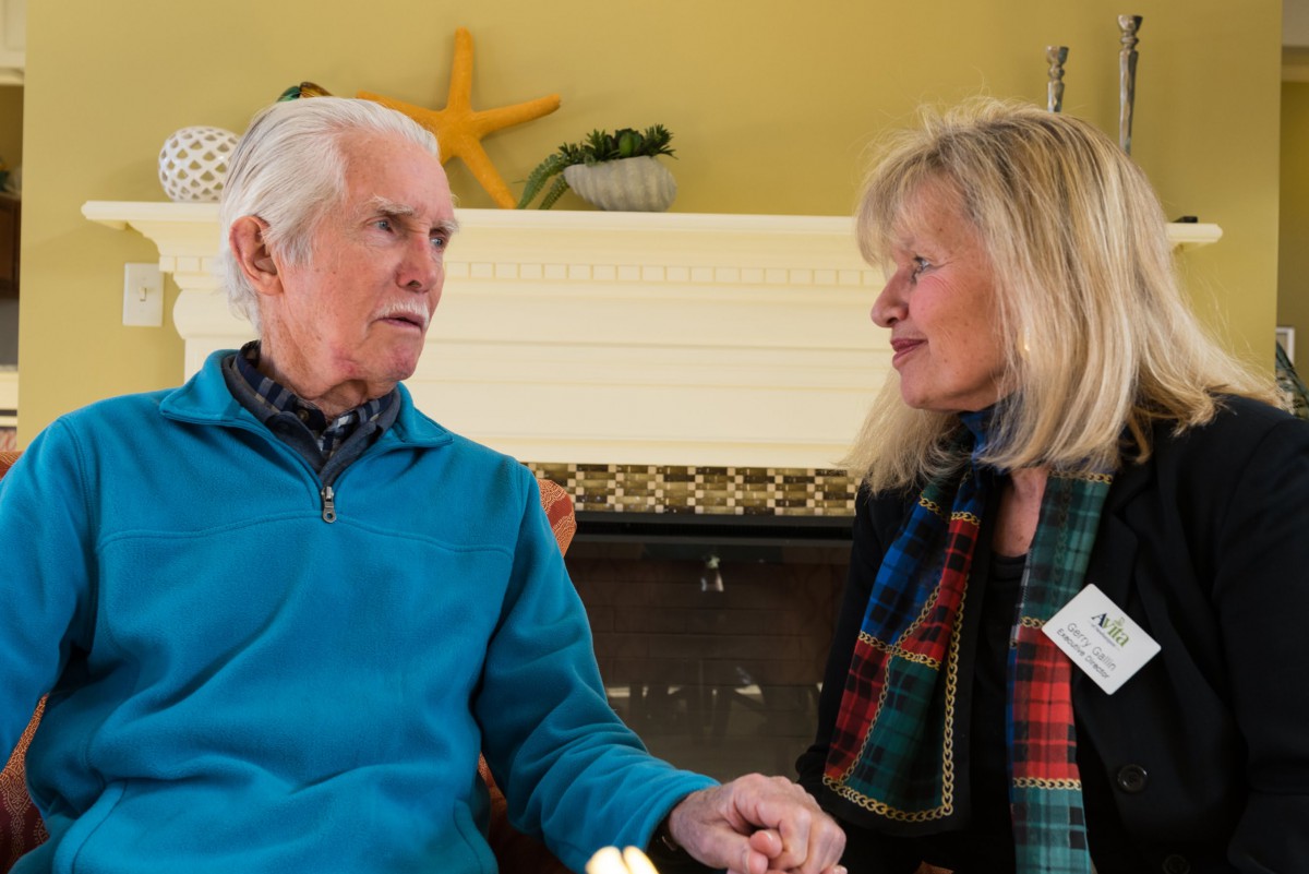 An elderly man and woman holding hands while engaged in conversation indoors.