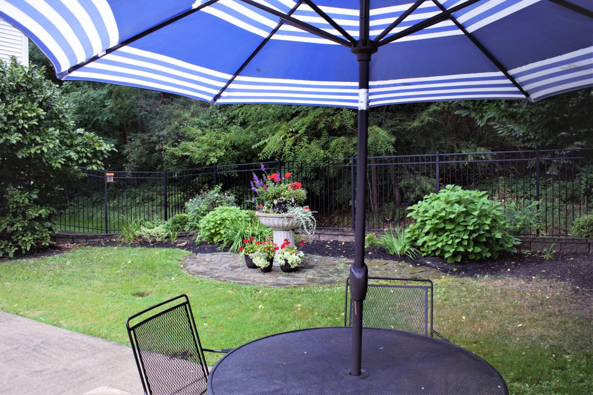A garden patio with a blue-striped umbrella, surrounded by lush greenery and decorative plants.