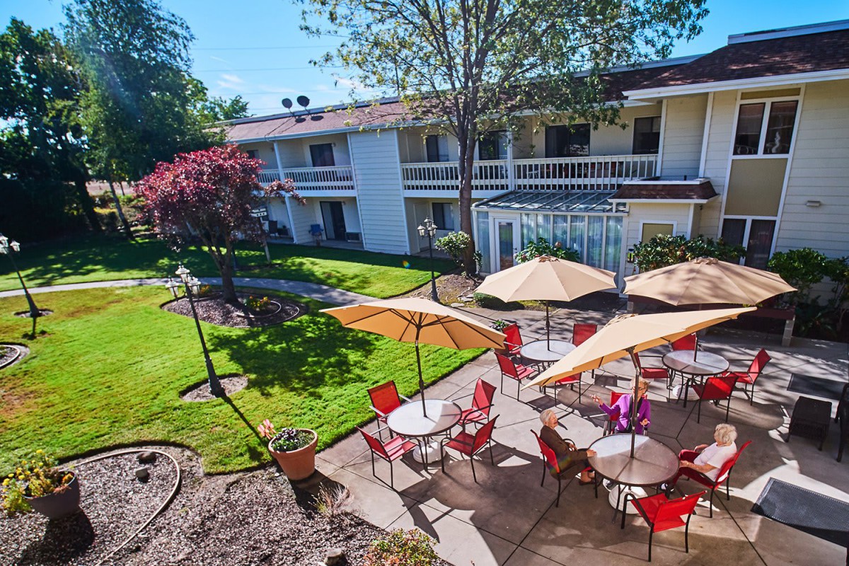 A sunny patio area with tables and umbrellas, surrounded by a landscaped garden and a two-story building in the background.
