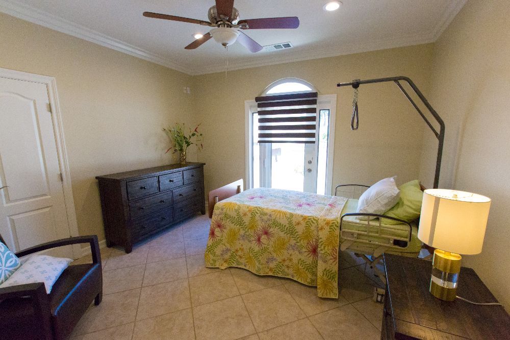 A cozy bedroom featuring a hospital bed near a window with striped blinds, alongside a dark dresser and beige tile flooring.