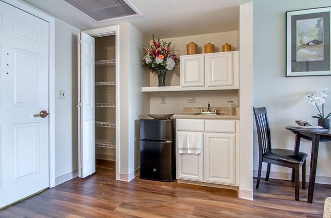 A small kitchenette with white cabinets, a mini fridge, and decorative flowers on display.