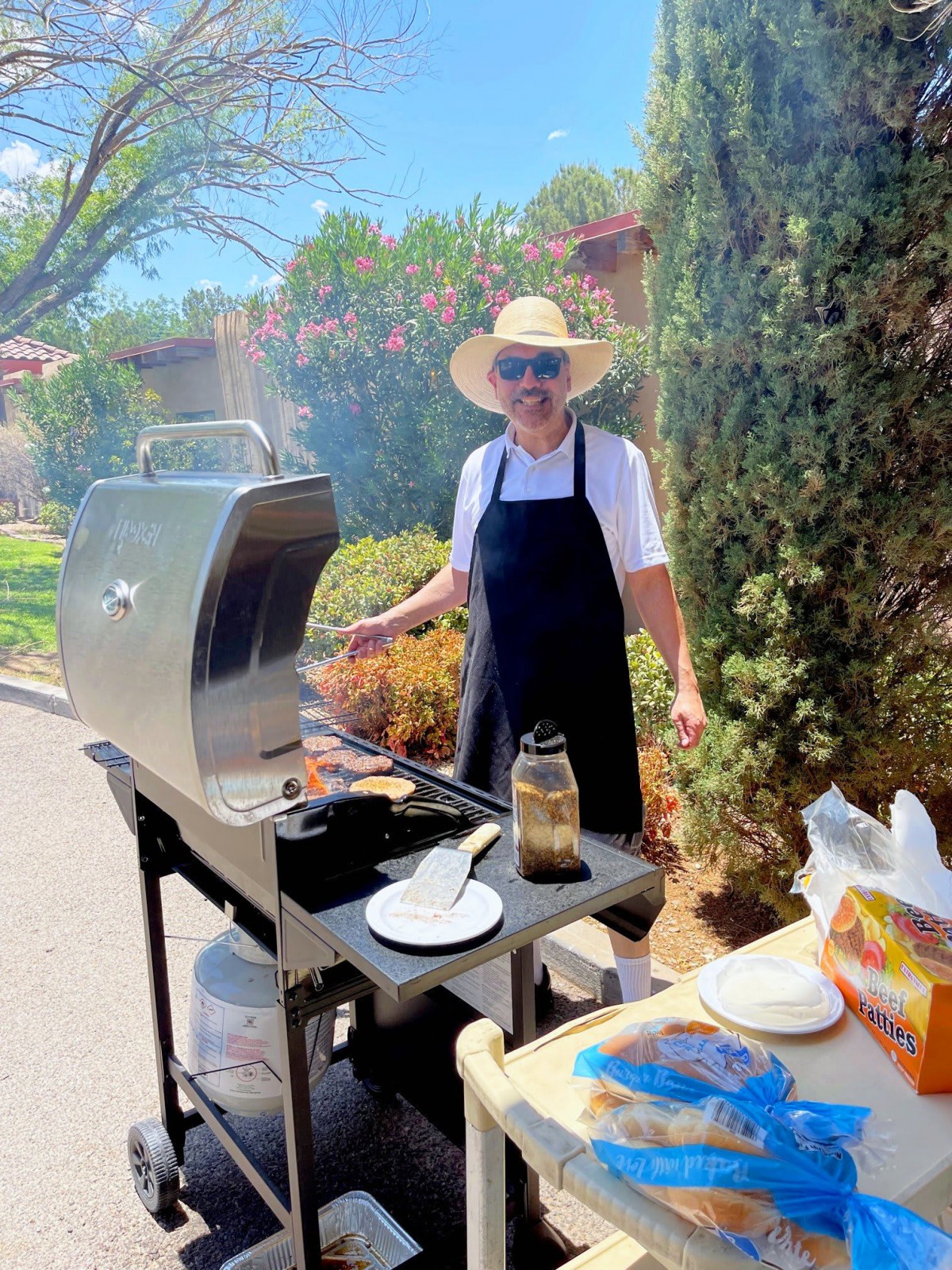 A person wearing a hat and apron is using a barbecue grill outdoors on a sunny day.
