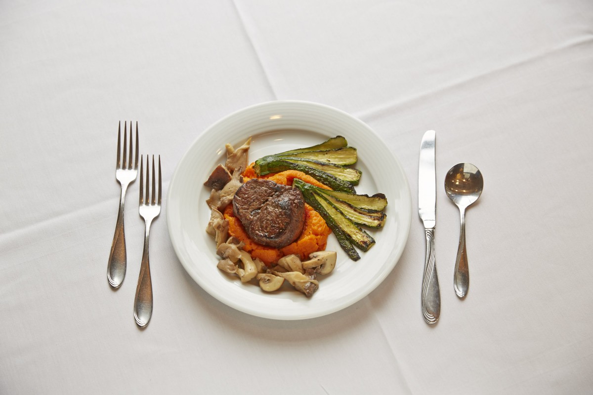 A gourmet meal with steak, vegetables, and pasta served on a white plate with cutlery arranged neatly on a table.