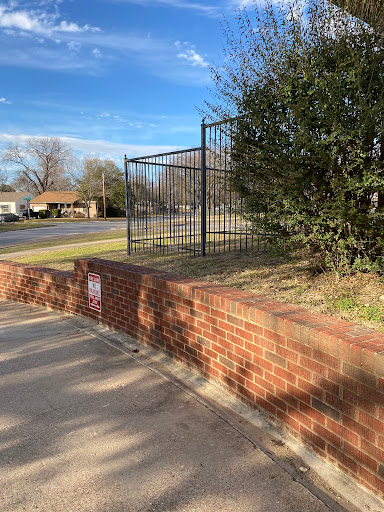 A view of a fenced area next to a brick wall, with grass and a clear blue sky.
