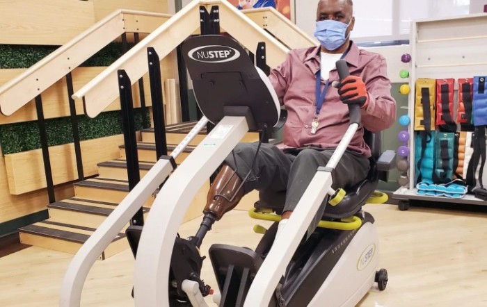 A person using adaptive exercise equipment during rehabilitation, wearing a mask and a prosthetic leg.