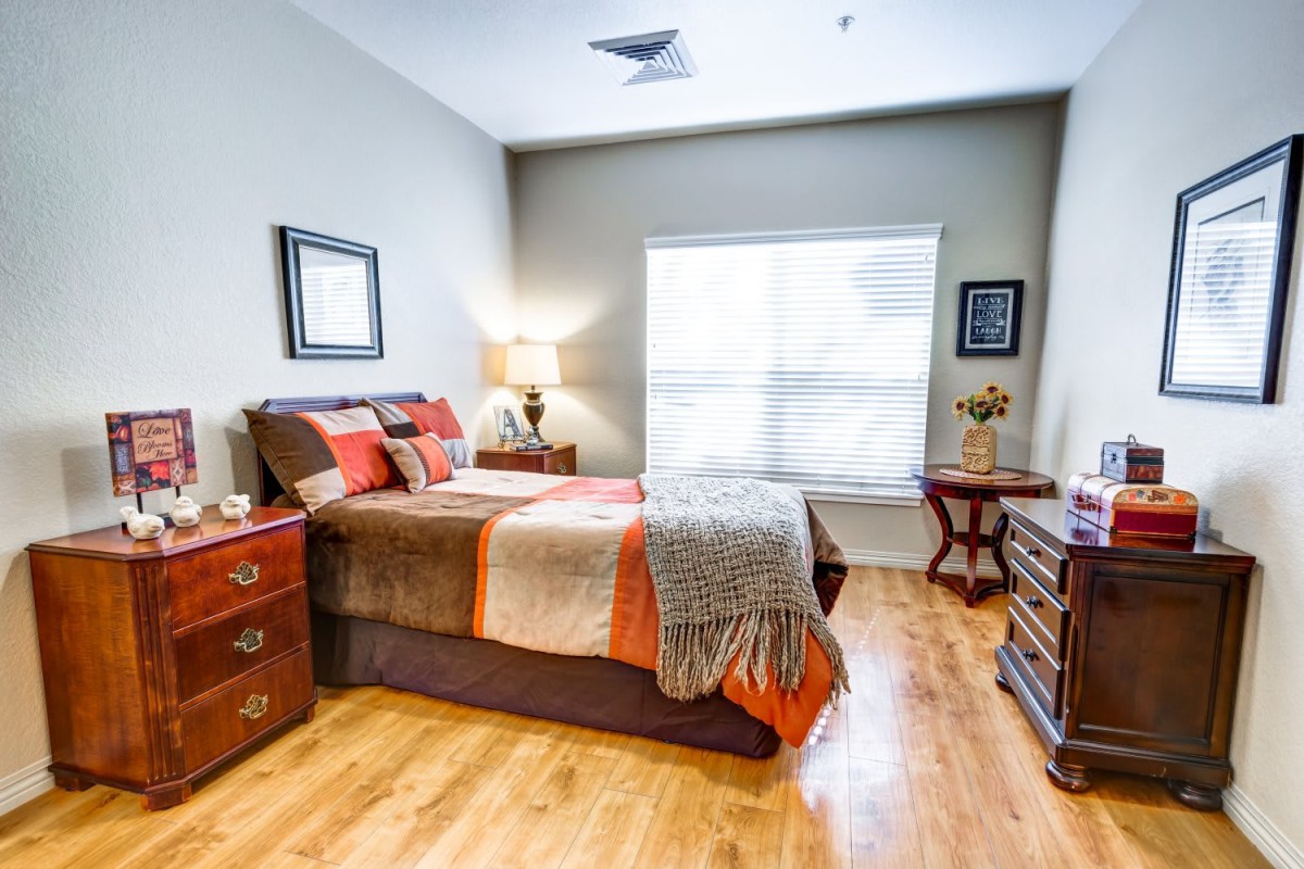 Cozy and well-decorated bedroom with a wooden floor, featuring a bed, nightstands with lamps, and a large window letting in natural light.
