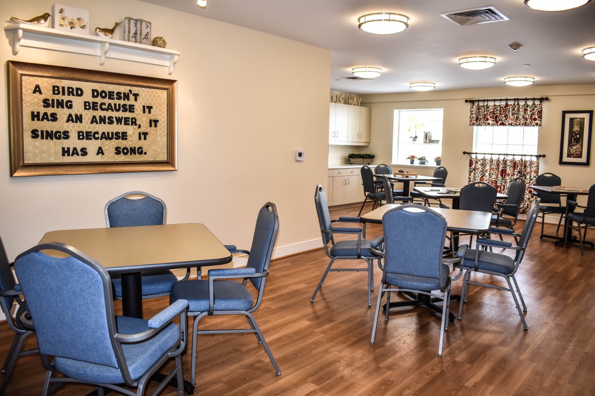 A cozy dining area with blue chairs, tables, and a small kitchen in the background.