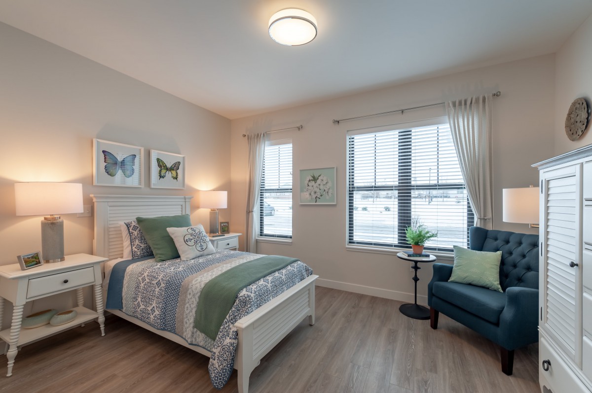 A cozy bedroom with a neatly made bed, artwork on the walls, and a comfortable chair by the window.