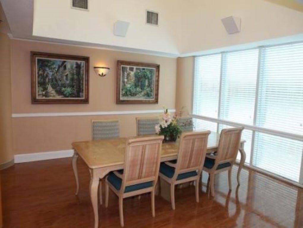 A well-lit dining room with a wooden dining table surrounded by six chairs, adorned with a vase of flowers. Artwork hangs on the wall, and large windows with blinds allow natural light to fill the space.