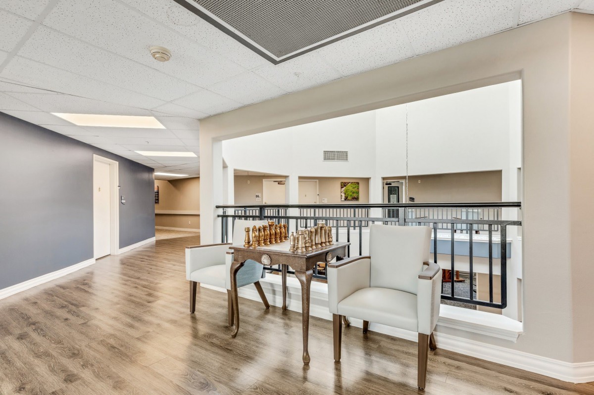 A well-lit interior space featuring a small table set with a chess board and two upholstered chairs on a wooden floor, adjacent to a railing overlooking a large area.