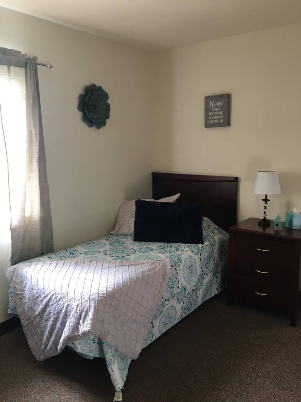 A cozy bedroom featuring a single bed with patterned bedding, a nightstand with a lamp, and wall decor.