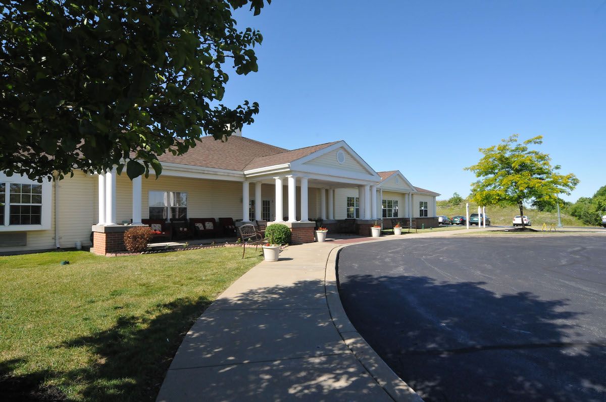 A large building with a columned entrance and a driveway, set in a landscaped area on a sunny day.