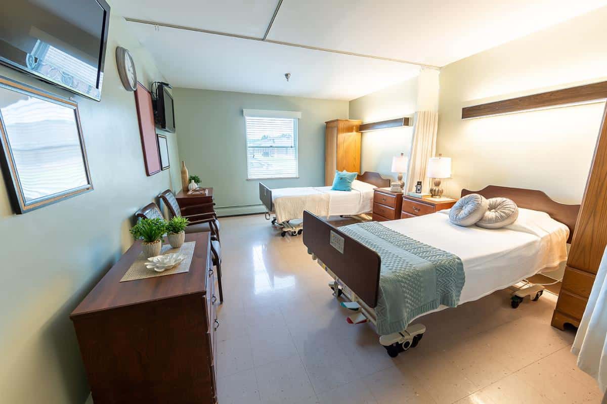 A well-lit hospital room featuring two patient beds with comfortable bedding and a window providing natural light.