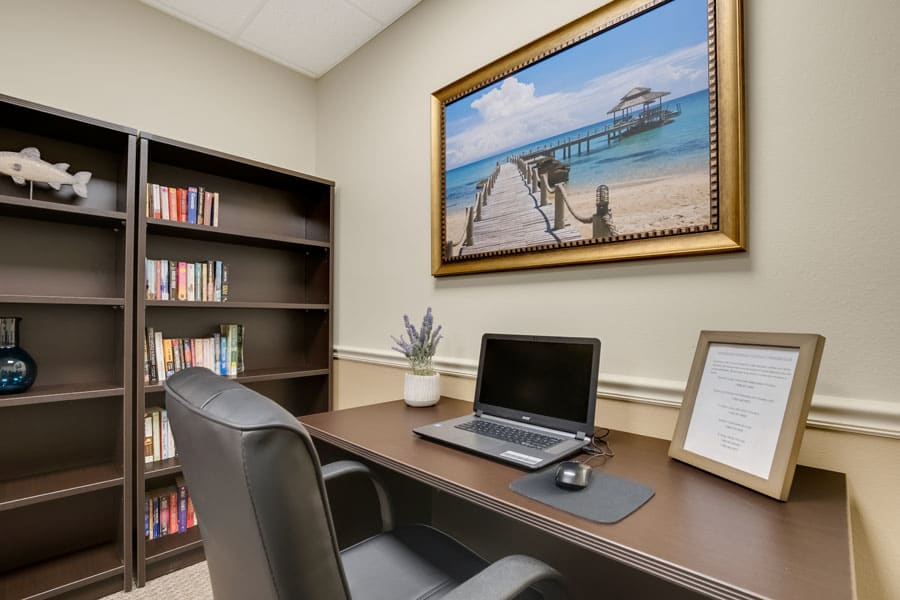 Modern office space with a desk and laptop, featuring a bookshelf and beach-themed painting.