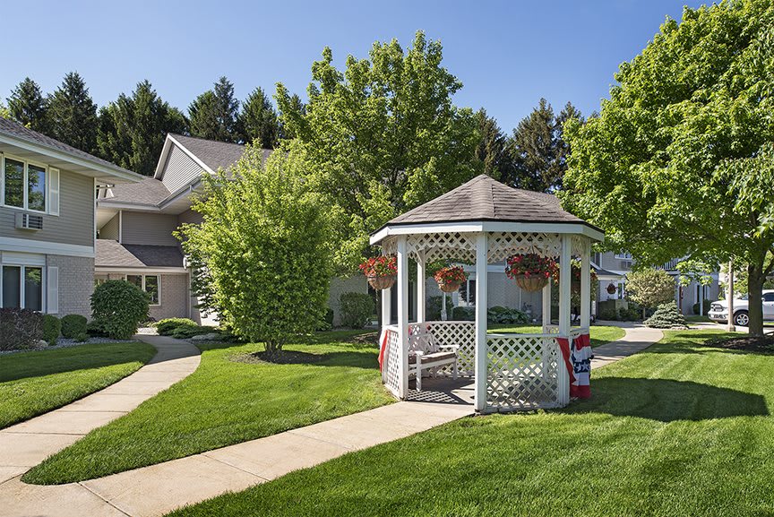 A scenic outdoor area featuring a white gazebo surrounded by lush green lawns and trees under a clear blue sky.