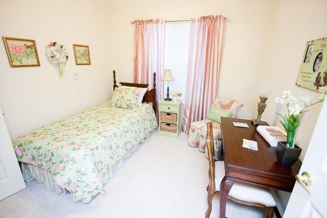 A cozy bedroom with a single bed featuring floral bedding, pink curtains, a desk with a chair, and a lamp on the side table.