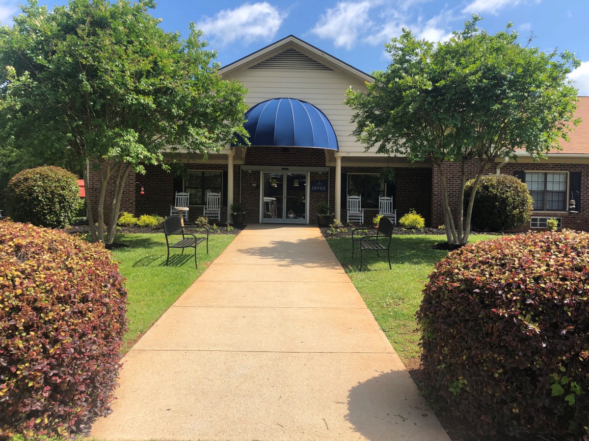 A walkway leads to a building entrance with a blue awning, surrounded by trees and neatly trimmed bushes.