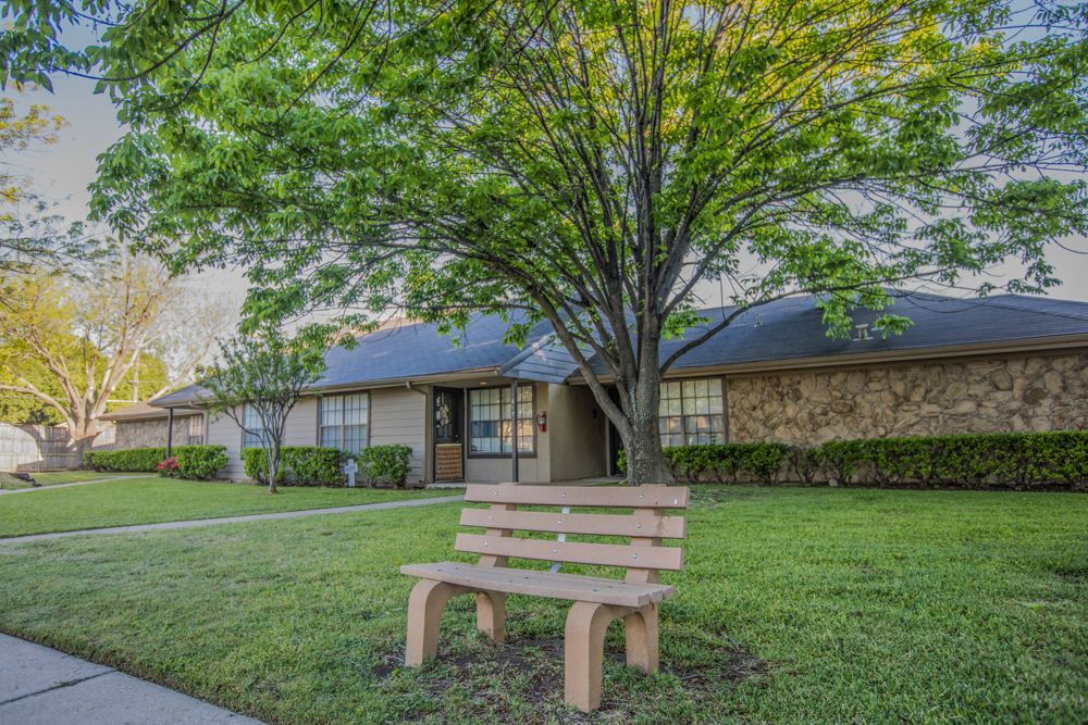 A suburban scene featuring a bench on a lush green lawn in front of a house with trees.