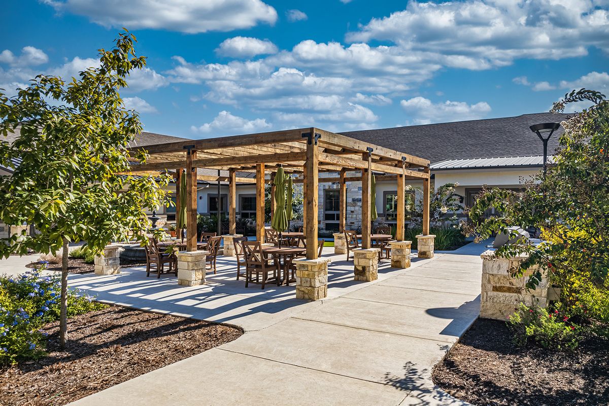 A sunny outdoor patio with a wooden pergola and seating area, surrounded by a landscaped garden.