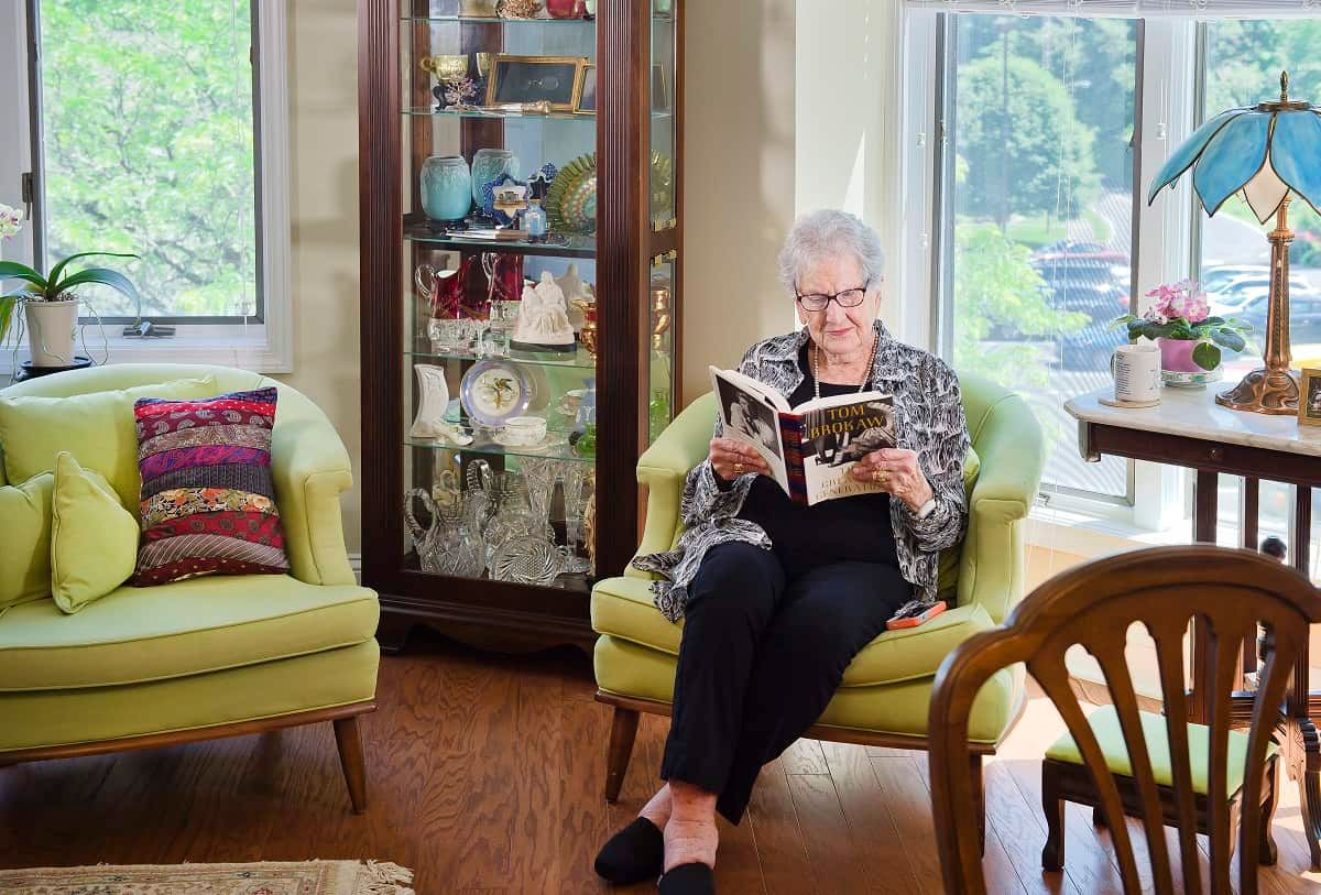An elderly woman sits comfortably in an armchair, reading a book in a cozy living room.
