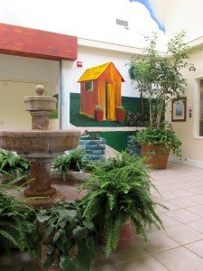 A serene indoor courtyard featuring a fountain surrounded by lush greenery and a colorful mural on the wall.