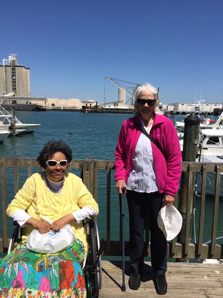 Two individuals enjoying a sunny day by the harbor, with boats docked in the background.