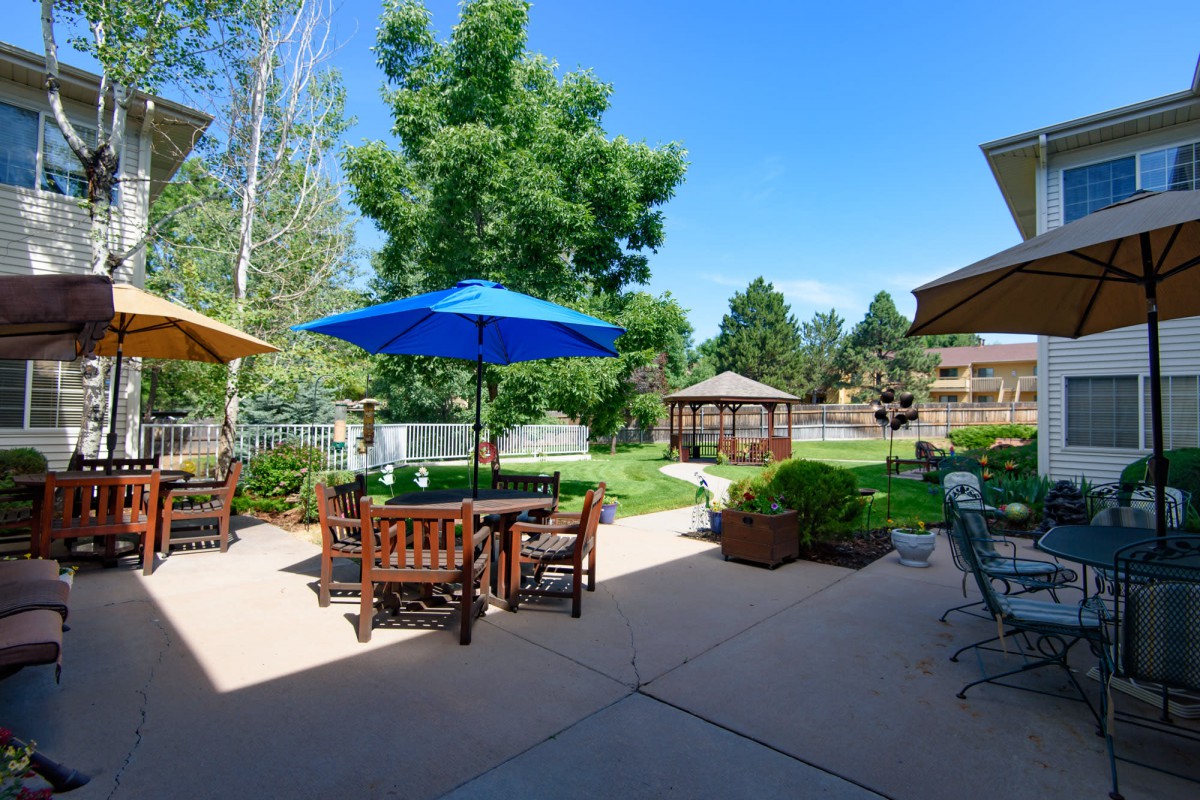 A scenic outdoor patio area with tables and chairs under umbrellas, surrounded by lush greenery and a gazebo.