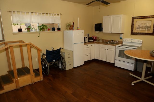 An accessible kitchen with a wheelchair, featuring white cabinets, appliances, and a wooden floor.