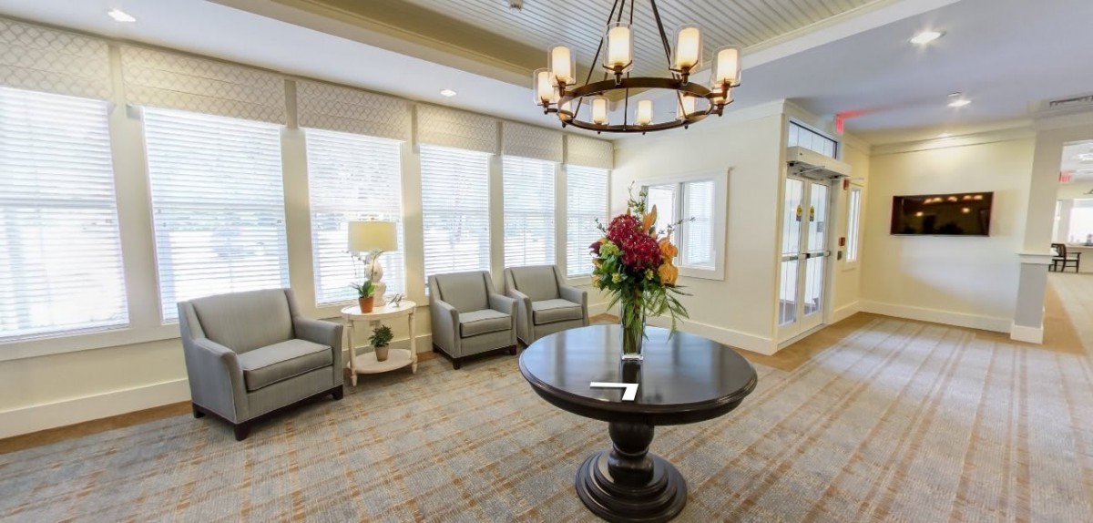 A well-lit lobby with comfortable seating, featuring a central round table adorned with a floral arrangement and an elegant chandelier overhead.
