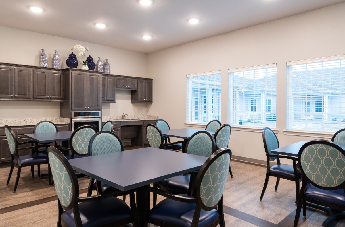 A well-lit dining area with multiple tables and chairs, featuring a kitchenette in the background.