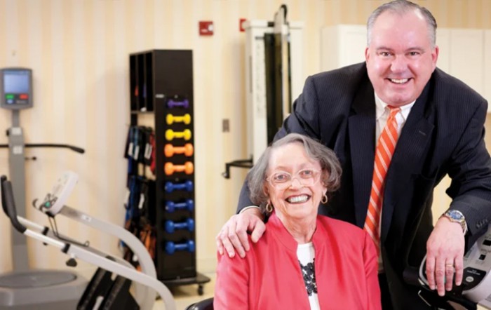 An elderly woman smiling with a man in a suit beside her, in a room with fitness equipment.