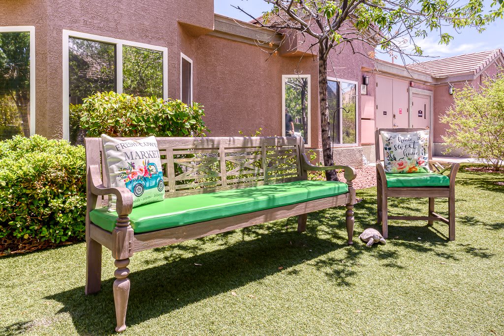 A sunny outdoor seating area with wooden benches adorned with green cushions, set in a backyard with a house and tree.