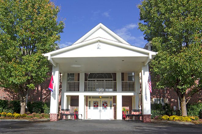 A building entrance with a white porch, flanked by trees and flags.