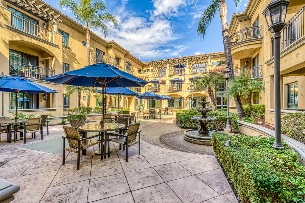 A sunny courtyard featuring a fountain, outdoor seating with blue umbrellas, surrounded by a multi-story building and palm trees.