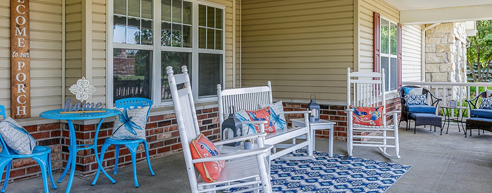 A cozy porch setup with white rocking chairs and a bench, decorated with colorful pillows and lanterns on a patterned rug.