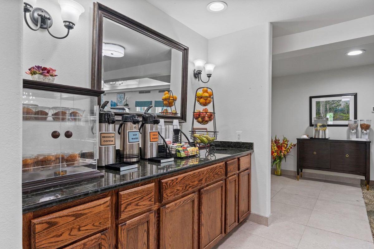 A cozy hotel breakfast area featuring a coffee station with beverage dispensers and assorted fruits on a granite countertop.