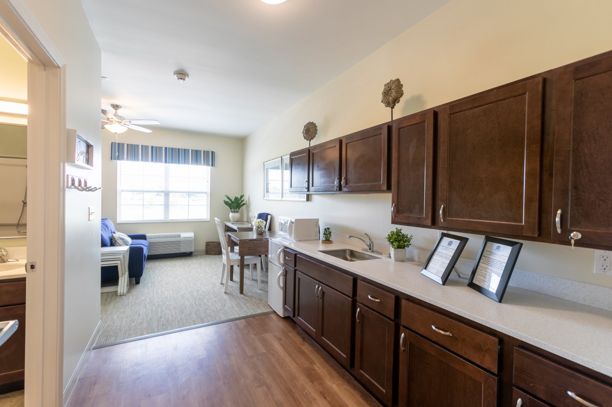 A modern kitchen and living area with wooden cabinets and flooring, featuring a dining table under bright natural light.