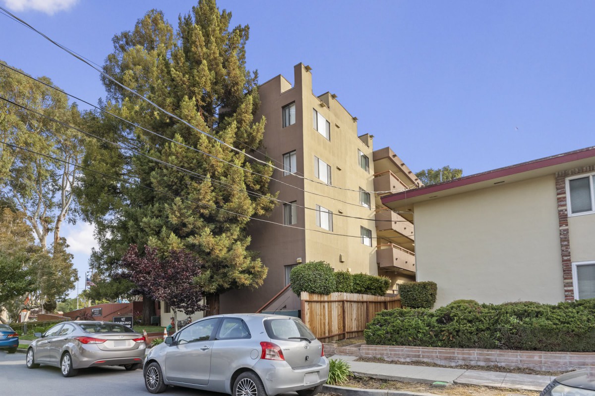 A residential street featuring an apartment building with parked cars and surrounding trees.