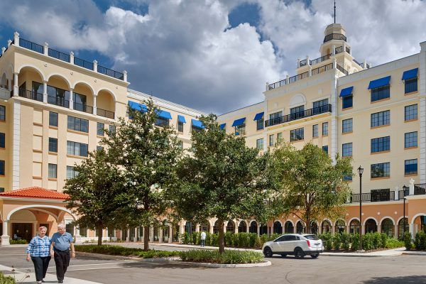A grand hotel building with arched windows and a decorative tower, set against a cloudy sky, surrounded by trees and parked cars.