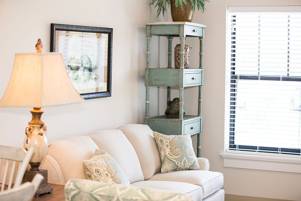 A cozy living room with a white sofa, decorative pillows, and a tall shelving unit with plants near a window.
