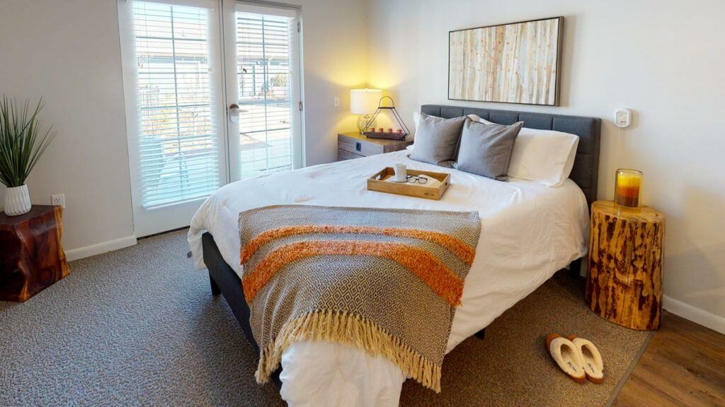 A cozy, well-decorated bedroom with a bed featuring a colorful blanket, pillows, and natural light streaming in through large windows.