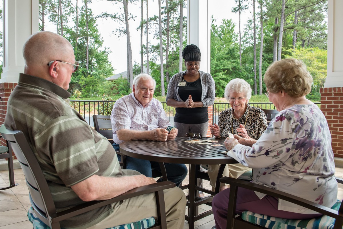A group of seniors playing cards on a patio with a staff member standing by.