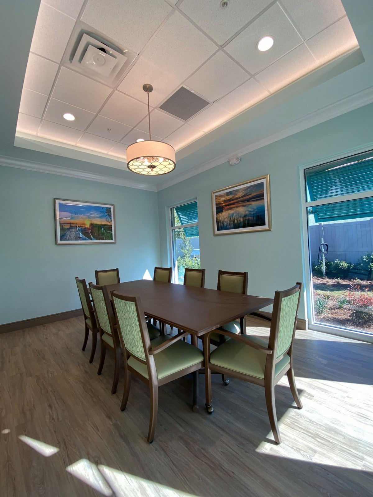 A modern dining or conference room with a large table, surrounded by eight chairs, featuring artwork on the walls and natural light from the windows.