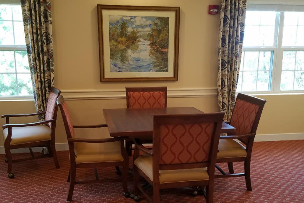A cozy dining area with a wooden table and chairs, adorned with a landscape painting and natural light from a window.
