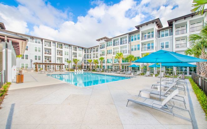A modern apartment complex with a large outdoor swimming pool and relaxation area on a sunny day.