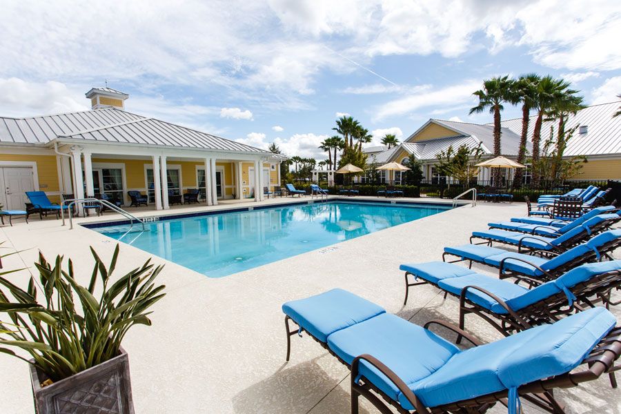 A serene outdoor swimming pool area with sun loungers, surrounded by palm trees and resort buildings under a clear blue sky.