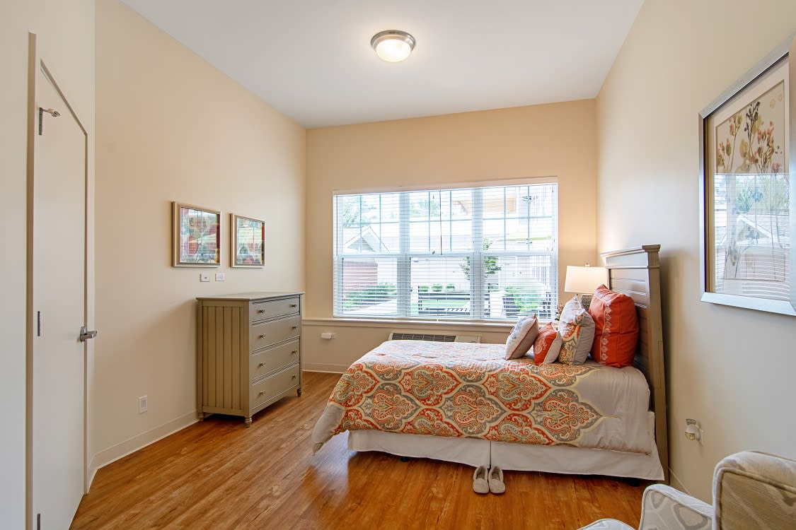 A cozy bedroom with a large window providing natural light, featuring a bed with patterned bedding and a chest of drawers.
