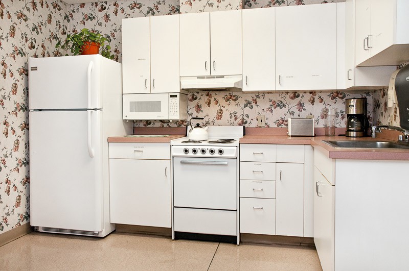 A retro-style kitchen featuring white cabinets, a stove, refrigerator, and microwave with floral wallpaper.