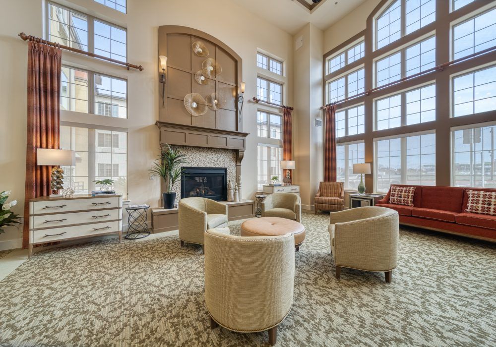 A spacious living room with a high ceiling, featuring large windows, a cozy seating area around a fireplace, and an elegant chandelier.