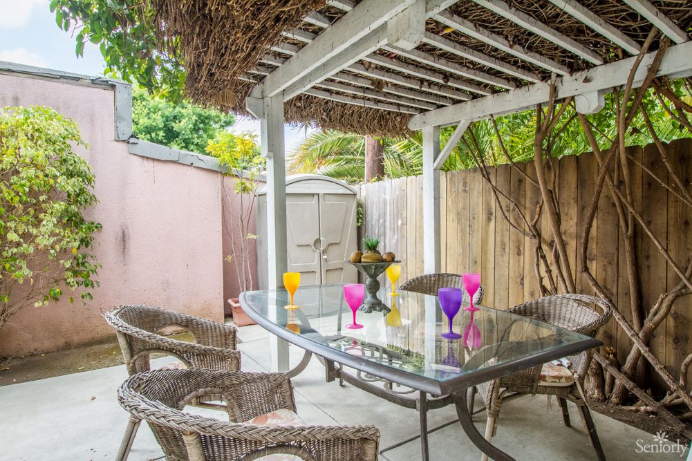 A cozy backyard patio with a pergola, wicker chairs, and a glass table adorned with colorful glasses.